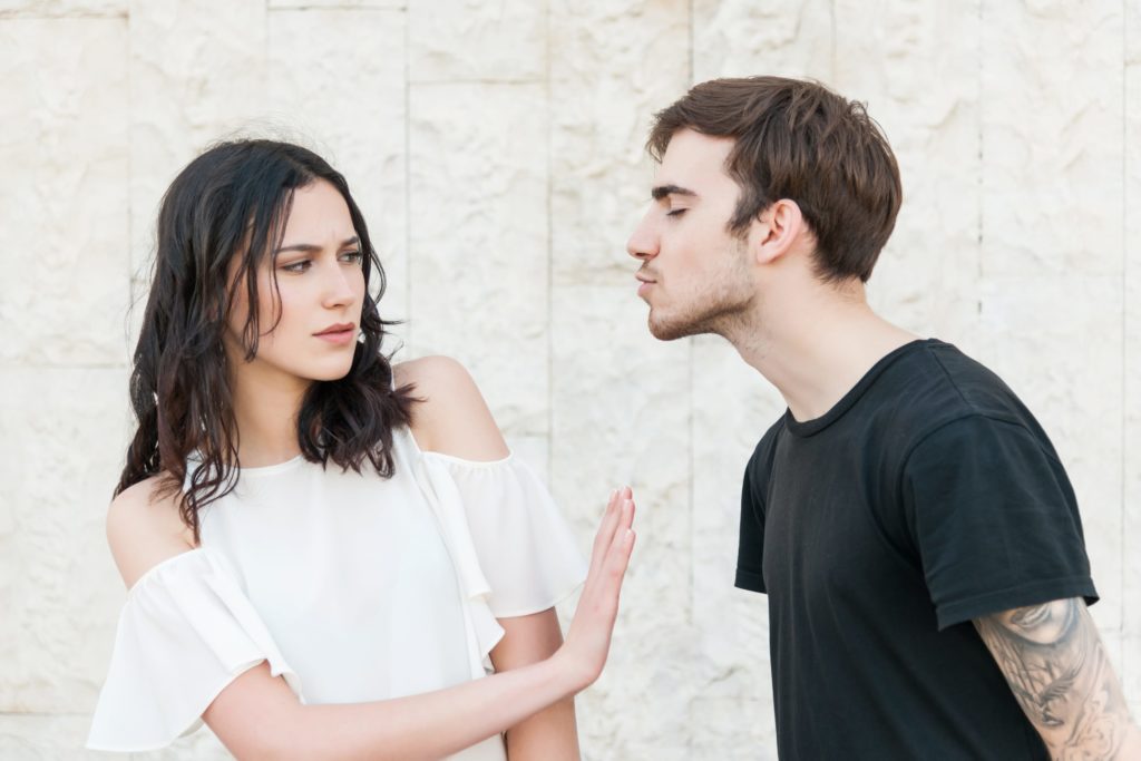 A young guy is leaning in to kiss a girl be she is frowning and making a hand gesture to stop him. They are wearing casual clothes.
