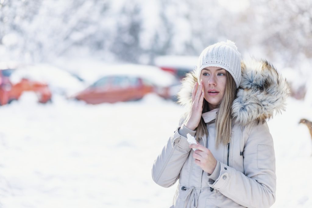 Portrait of a beautiful woman in snow with application of the protective cream in winter. Portrait of a beauty happy lady applying facial moisturiser cream in winter